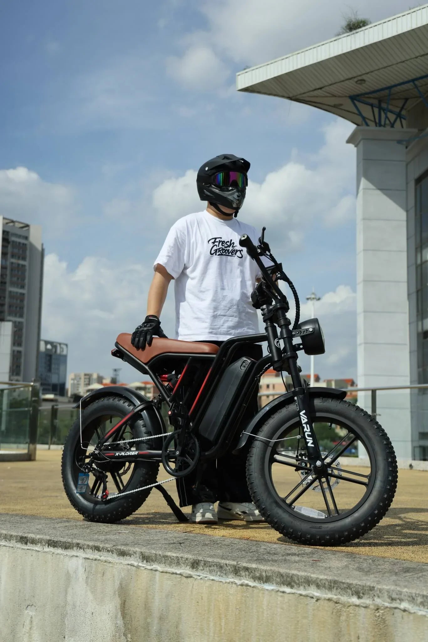 Person in white Fresh Groovers t-shirt and black helmet standing with black Varun electric bike outdoors