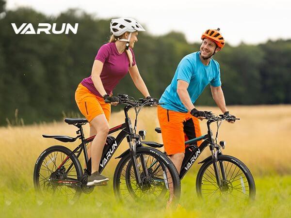 Man and woman in helmets riding Varun electric bikes on grassy field with forest background