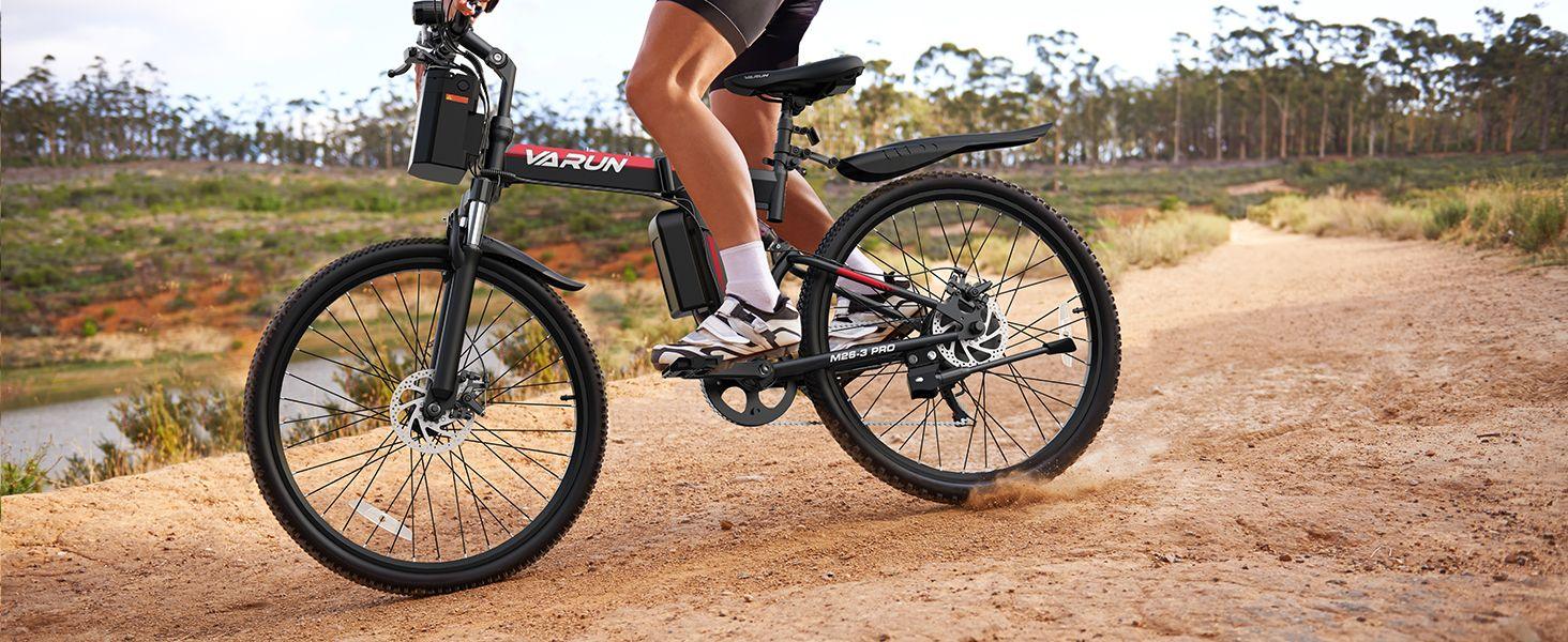 Person riding black Varun M26-3 Pro electric mountain bike on dirt trail in natural outdoor setting