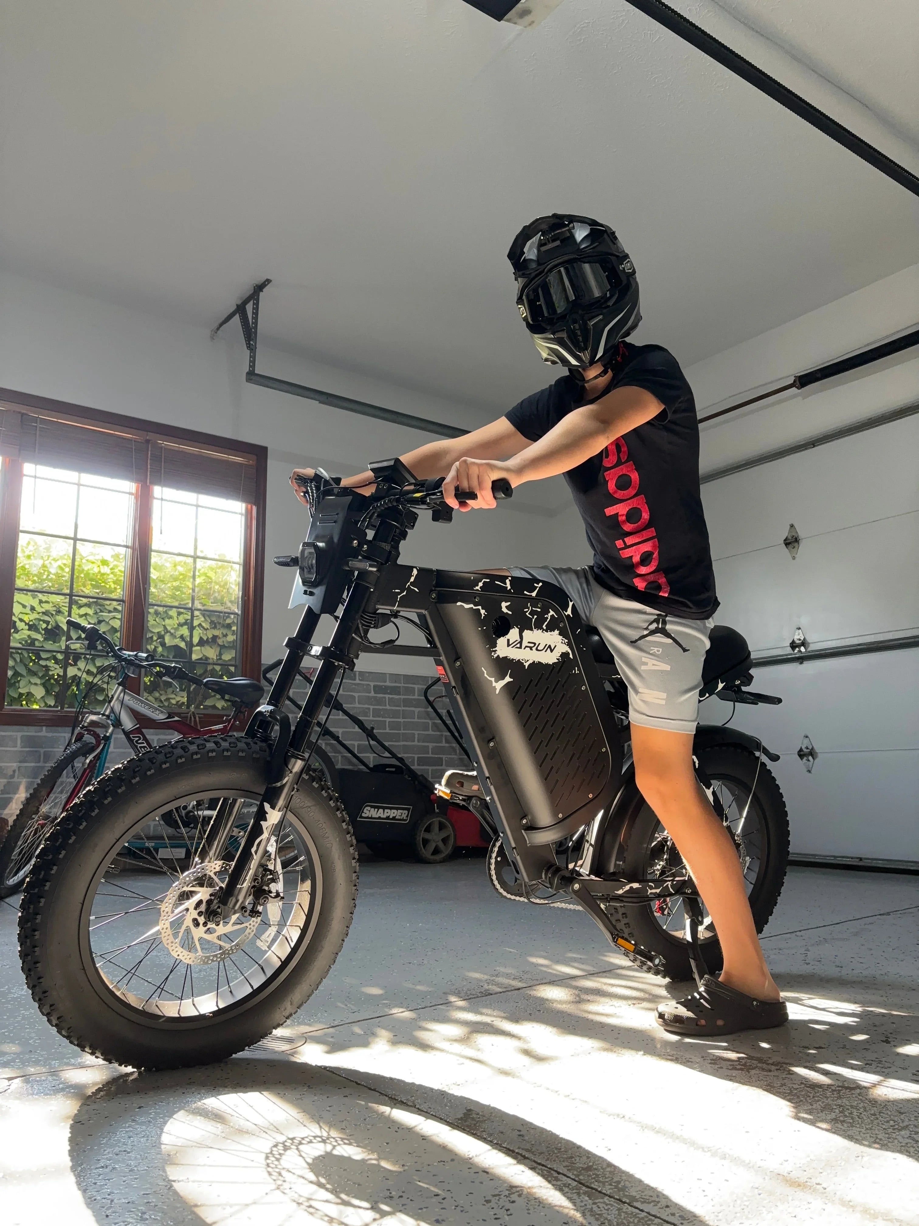 Person wearing black helmet and Adidas shirt sitting on black Varun electric bike inside garage
