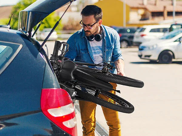 Man loading a black mountain bike into the trunk of a black hatchback car in a parking lot