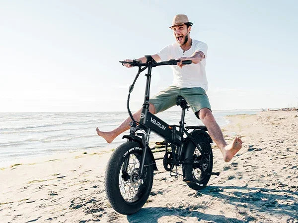 Man joyfully riding a black Varun electric bike barefoot on a sunny beach with clear sky