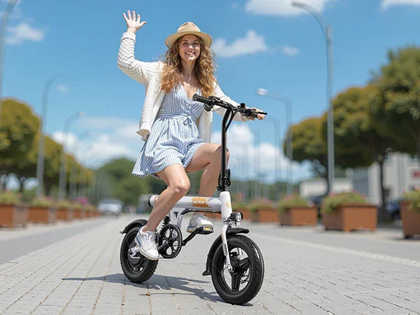 young woman wearing hat riding compact electric bike on sunny paved street waving hand