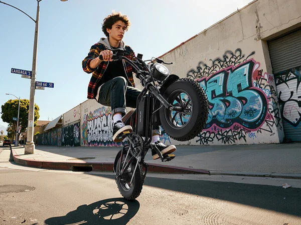 Young man performing a wheelie on electric bike near graffiti-covered wall on urban street