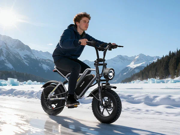 Young man riding black electric bike on icy lake with snowy mountains and blue sky background