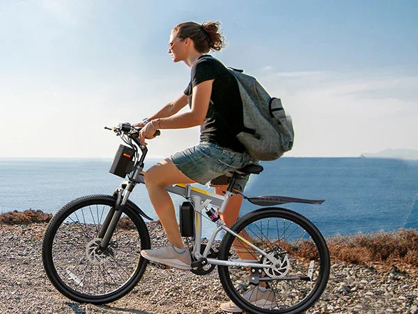 Woman riding electric mountain bike along rocky coastline with ocean backdrop, wearing casual summer clothes and backpack