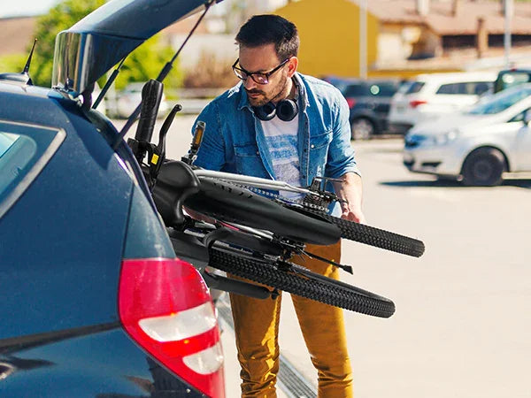 Man loading a folded black bicycle into the trunk of a blue car in a sunny parking lot