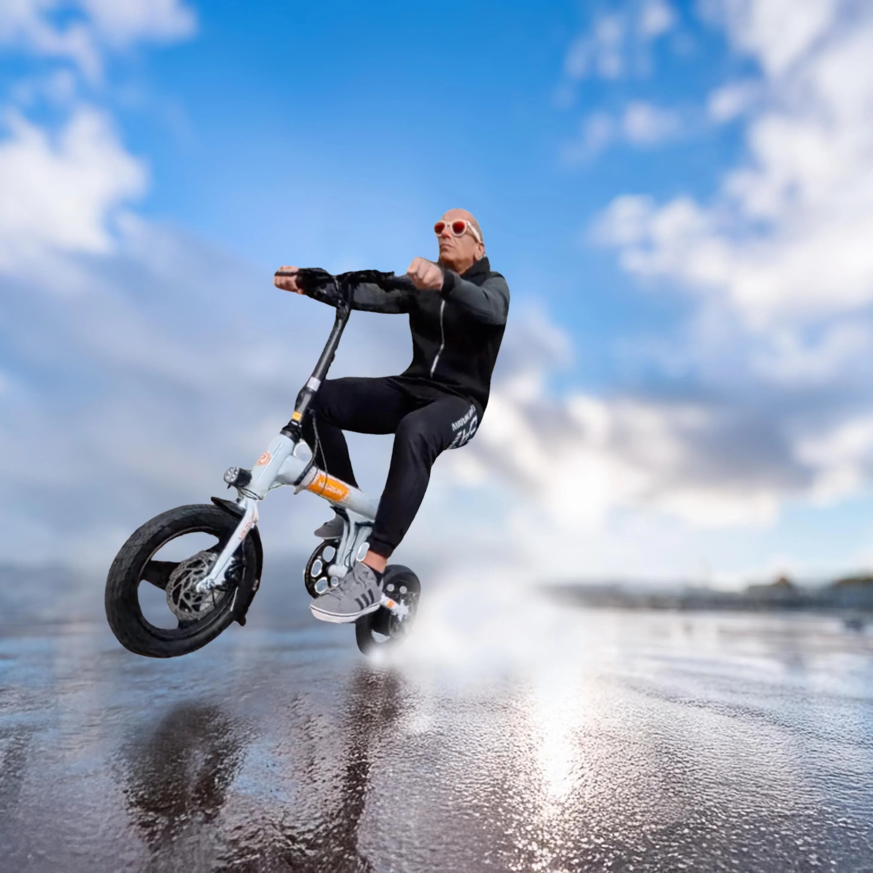 Man in black outfit and red goggles riding a small electric bike on a wet reflective surface under blue sky