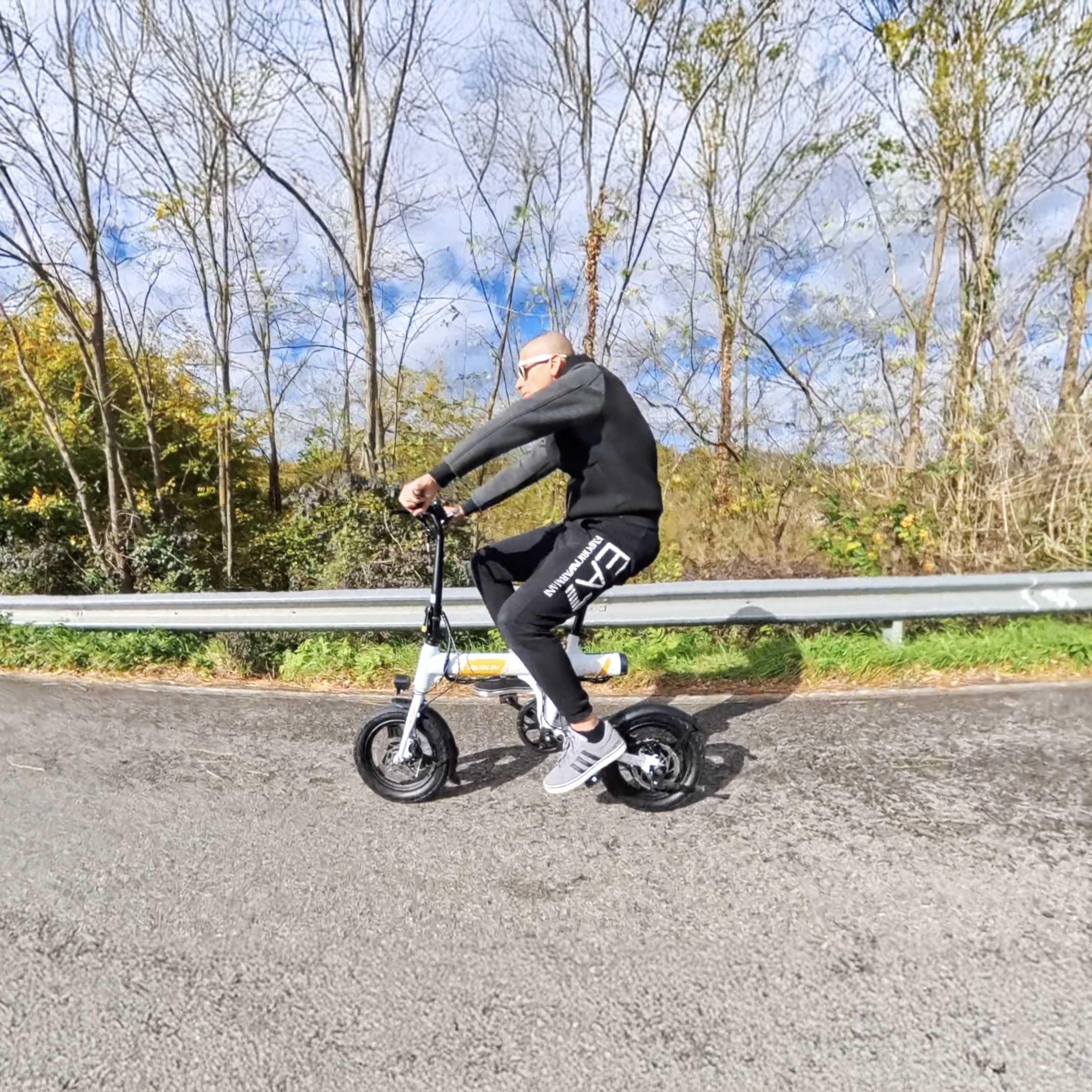 Bald man in black sportswear riding a compact white electric bike on a road with trees and guardrail
