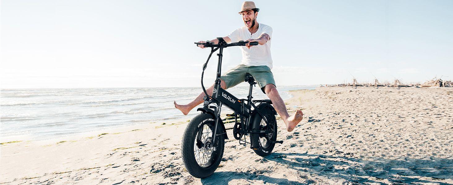 Man wearing a hat and shorts riding a black Varun electric bike barefoot on a sandy beach.