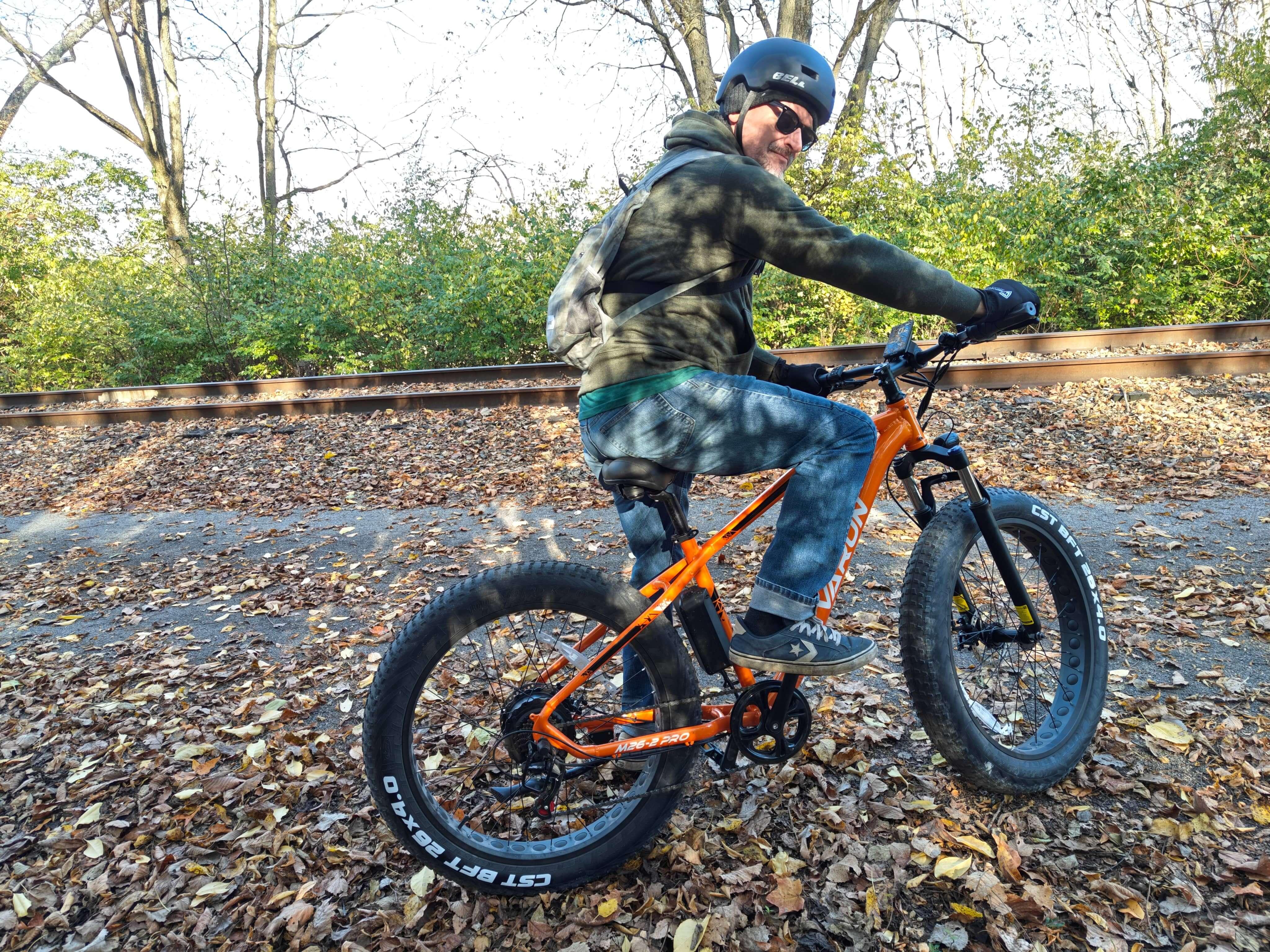 Man wearing helmet and sunglasses riding an orange fat tire electric bike on leafy trail near railroad tracks