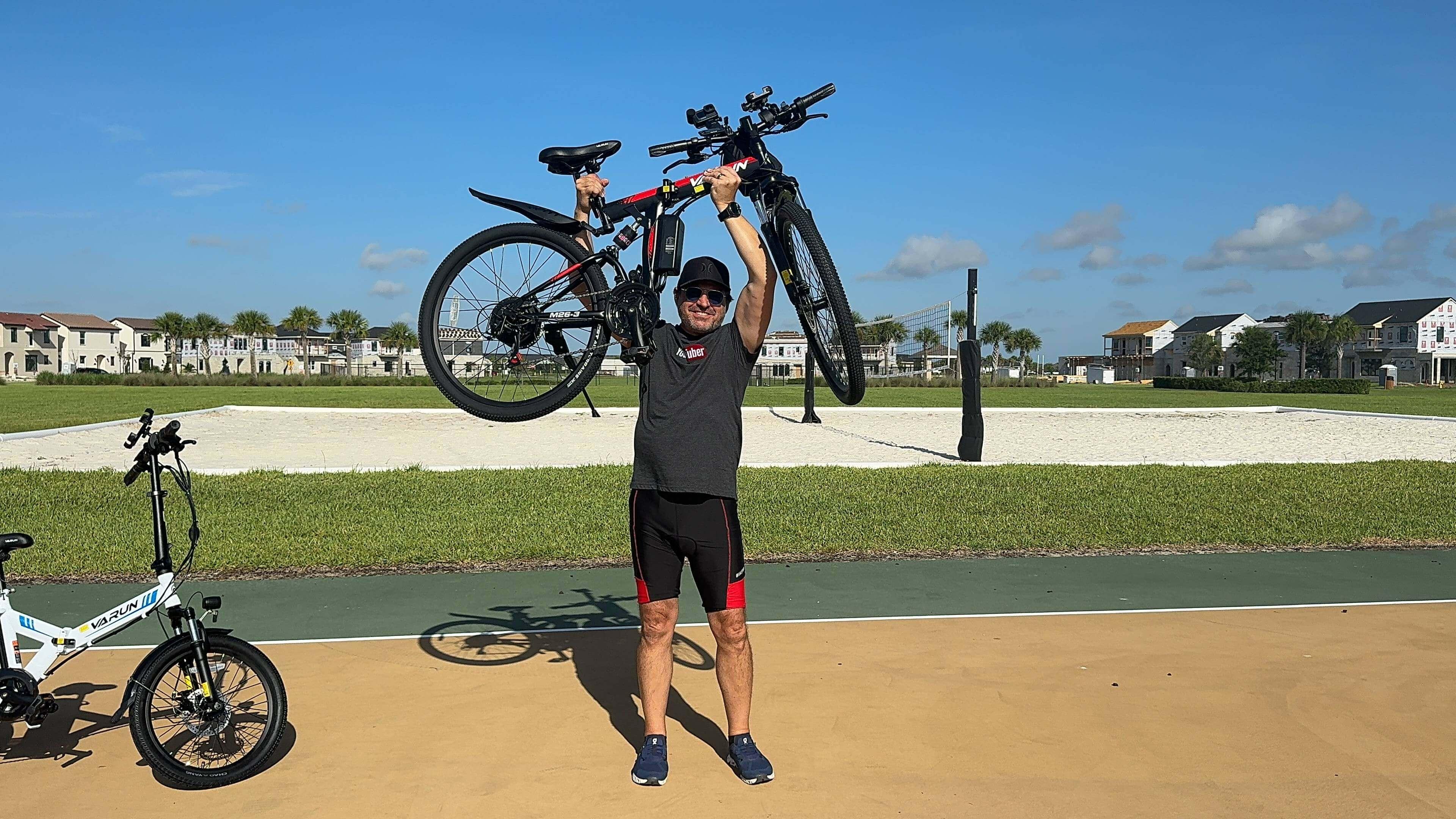Man lifting black mountain bike on court with volleyball net and houses in background