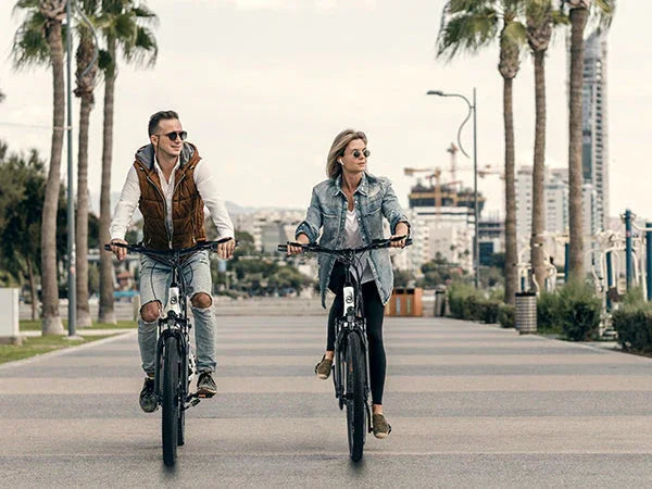 Couple riding electric bikes on palm-lined urban path with city buildings in background