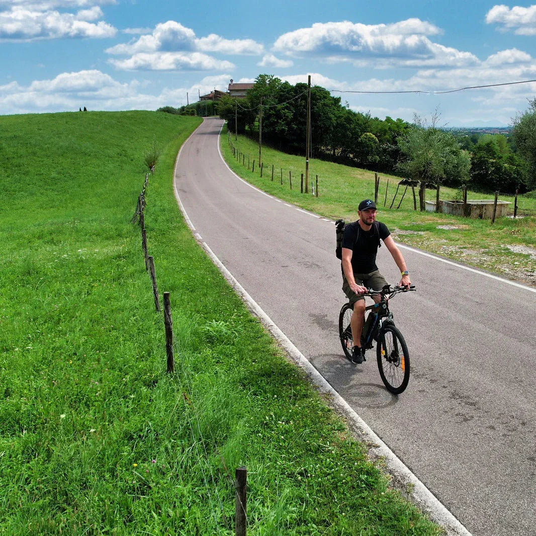 Man riding electric bike on rural road surrounded by green fields and trees under blue sky