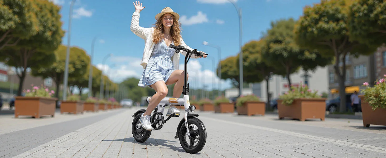 Young woman in hat and dress riding electric bike on sunny city street waving