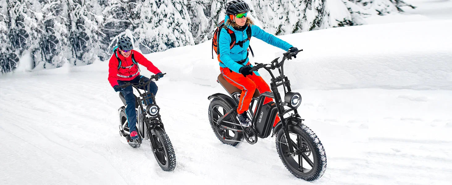 Two people riding black fat-tire electric bikes on a snowy trail with snow-covered trees
