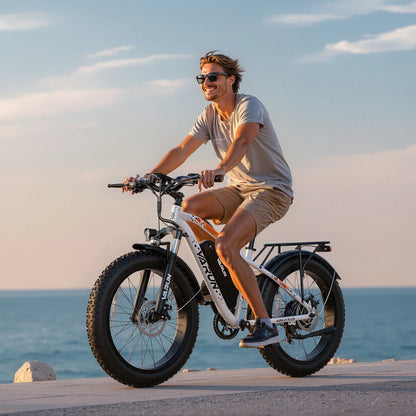 Smiling man wearing sunglasses riding a white Varun electric fat tire bike by the sea at sunset